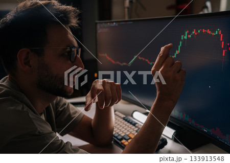Positive young man smiling and pointing at fluctuating financial graph on desktop screen, actively managing cryptocurrency investments and tracking market trends for profitable trading decisions. Positive young man smiling and pointing at fluctuating financial graph on desktop screen, actively managing cryptocurrency investments and tracking market trends for profitable trading decisions. 133913548