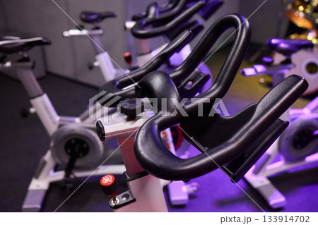 Cycle gym gear, Closeup of exercise bike with purple lighting and textured grips, Detailed view of spin bike handlebars and surroundings emphasizing fitness environment Cycle gym gear, Closeup of exercise bike with purple lighting and textured grips, Detailed view of spin bike handlebars and surroundings emphasizing fitness environment 133914702