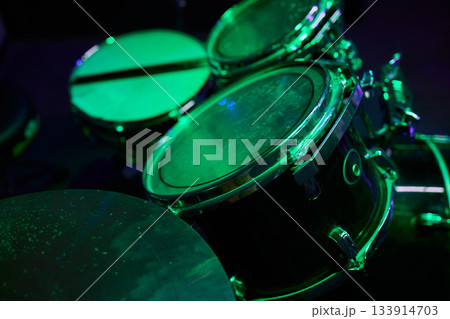 Greenlit drum toms close-up showing textured heads, reflective rims and subtle dust under moody stage lighting, empty kit poised for band rehearsal and soundcheck Greenlit drum toms close-up showing textured heads, reflective rims and subtle dust under moody stage lighting, empty kit poised for band rehearsal and soundcheck 133914703