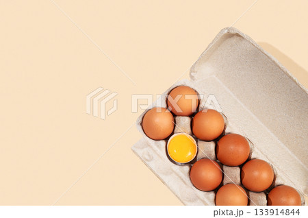 Fresh brown eggs in an open carton with one cracked yolk on a beige background Fresh brown eggs in an open carton with one cracked yolk on a beige background 133914844