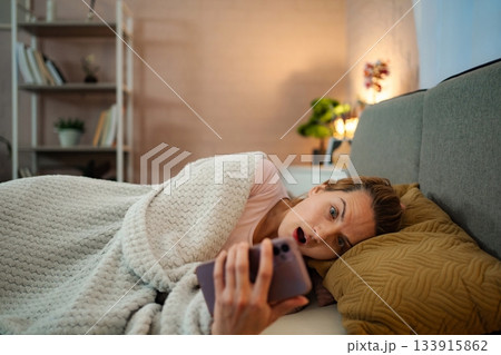 Woman doomscrolling on phone in bed looking shocked at news 133915862