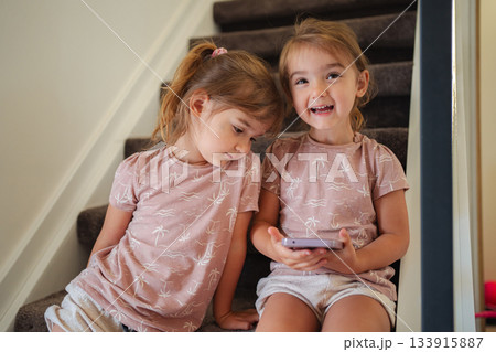 Two little sisters watching a smartphone sitting on stairs at home Two little sisters watching a smartphone sitting on stairs at home 133915887