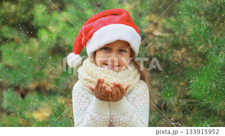 Christmas portrait of happy smiling little girl child in santa red hat blows snow in snowy forest 133915952