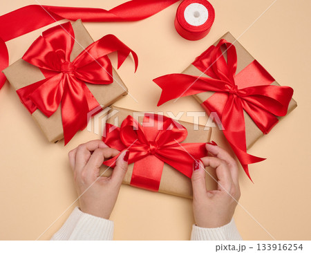 A woman's hands in a white sweater hold a gift box wrapped in craft paper with a red satin bow and a roll of ribbon. A woman's hands in a white sweater hold a gift box wrapped in craft paper with a red satin bow and a roll of ribbon. 133916254