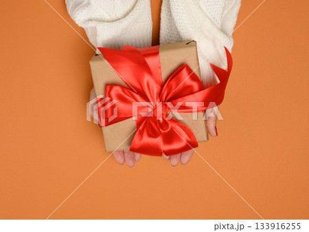 A woman's hands, wearing a white knitted sweater, hold a square gift box with a large red satin bow. 133916255