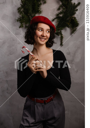 Caucasian woman holding candy cane in festive room with grey wall and green garland Caucasian woman holding candy cane in festive room with grey wall and green garland 133916409