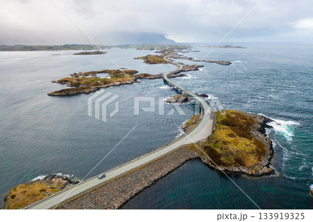Cars drive on Atlantic ocean road with bridge connecting large islands. Small house adds touch of human presence to dramatic Norwegian coastal scene 133919325