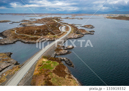 Atlantic ocean road demonstrates impressive span stretching across rocky islets in sea. Highway elegantly curves along path under muted cloudy sky Atlantic ocean road demonstrates impressive span stretching across rocky islets in sea. Highway elegantly curves along path under muted cloudy sky 133919332