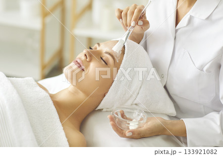 Female cosmetologist applying moisturizing mask with brush to a young woman in spa salon. Female cosmetologist applying moisturizing mask with brush to a young woman in spa salon. 133921082