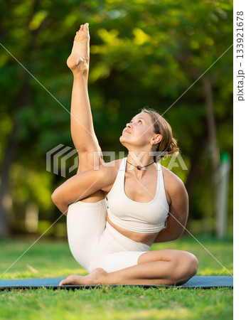 Girl in park on green grass of lawn performs Parivritta Surya Yantrasana 133921678