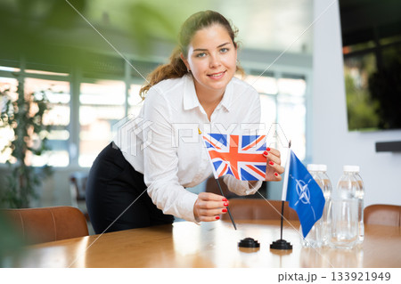 Woman secretary prepares an office for negotiations - she places flags of NATO (OTAN) and Great Britain table 133921949