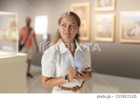Young girl holding an information booklet looks at paintings in an art exhibition in museum Young girl holding an information booklet looks at paintings in an art exhibition in museum 133922120
