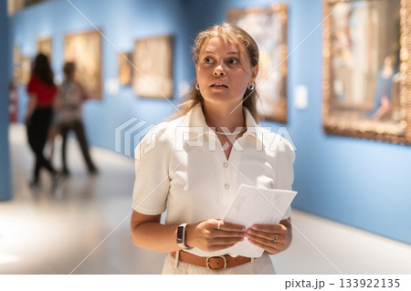 Portrait of young girl visitor with paper guide at the exhibition of paintings in museum 133922135