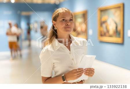 Woman with sheet piece of paper in hands view pictures and icons in museum hall Woman with sheet piece of paper in hands view pictures and icons in museum hall 133922138