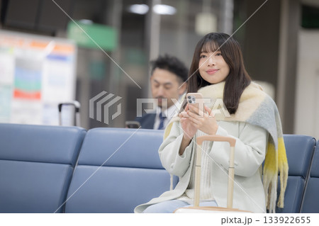 冬の空港ロビーでスーツケースを持つ若い女性の旅行シーン　ロビーで搭乗を待つ座る	 133922655