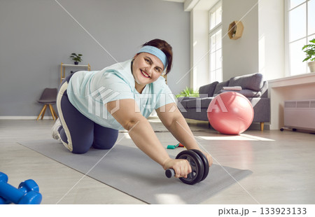 Happy, overweight woman doing fitness exercise with ab roller during her sports workout at home Happy, overweight woman doing fitness exercise with ab roller during her sports workout at home 133923133