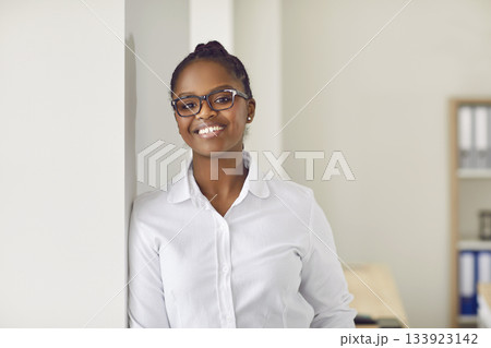Portrait of happy businesswoman standing by office window, looking at camera and smiling 133923142