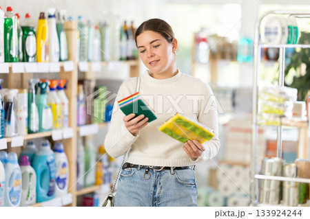 Young woman housewife in household store examines package, chooses abrasive sponge 133924244