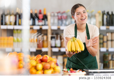 Young woman seller in store is standing near fruit counter, selling bananas. Young woman seller in store is standing near fruit counter, selling bananas. 133924308