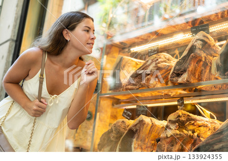 Passerby girl examines dried beef, aged meat, and quarters of carcass in shop window. Passerby girl examines dried beef, aged meat, and quarters of carcass in shop window. 133924355