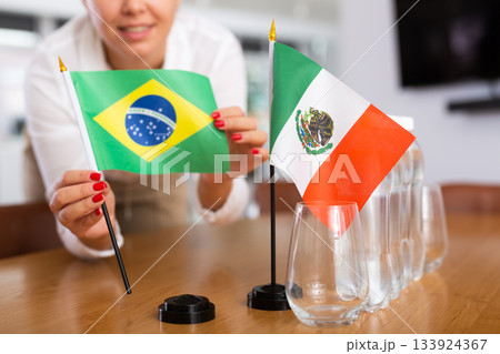 A woman in a white blouse with friendly smile arrangs flags of Mexico and Brazil for meeting. A woman in a white blouse with friendly smile arrangs flags of Mexico and Brazil for meeting. 133924367