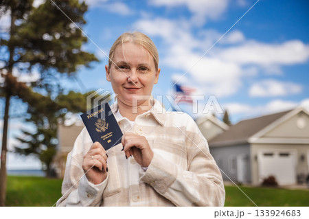 Smiling woman holding a United States passport outdoors with an American flag in the background, symbolizing citizenship, immigration and travel freedom. Smiling woman holding a United States passport outdoors with an American flag in the background, symbolizing citizenship, immigration and travel freedom. 133924683