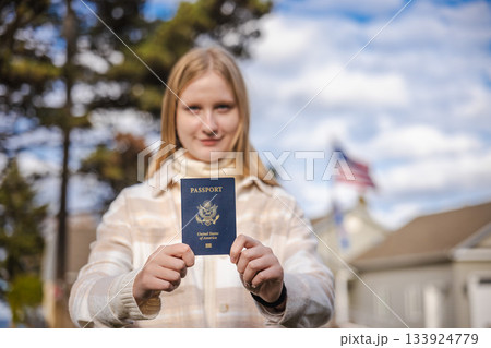 Young woman standing outdoors, extending her hands forward and holding a blue US passport, with suburban houses and an American flag visible in the background. Young woman standing outdoors, extending her hands forward and holding a blue US passport, with suburban houses and an American flag visible in the background. 133924779