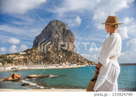 Young woman in a white summer dress standing by the sea in Calpe, Spain, looking toward the Penon de Ifach rock on a sunny day Young woman in a white summer dress standing by the sea in Calpe, Spain, looking toward the Penon de Ifach rock on a sunny day 133924790