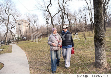 Two Russian friends aged 50 and 60 walk in Nagorny Park Vladivostok, enjoying calm movement and warm conversation that supports wellbeing and positive connection. 133927803