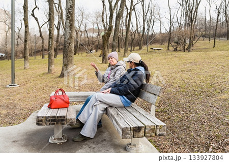 Two Russian friends aged 50 and 60 sit in Nagorny Park Vladivostok, enjoying calm spring conversation that supports emotional balance, connection and wellbeing. 133927804