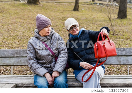 Two Russian friends 50 and 60 years old sit in a spring park as one proudly shows her new bag sharing calm joy mindful connection and shopping confidence. Two Russian friends 50 and 60 years old sit in a spring park as one proudly shows her new bag sharing calm joy mindful connection and shopping confidence. 133927805