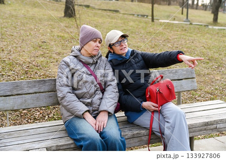 Two Russian friends 50 and 60 years old sit in a spring park as one points into the distance sharing calm presence curiosity and a relaxed thoughtful moment together. Two Russian friends 50 and 60 years old sit in a spring park as one points into the distance sharing calm presence curiosity and a relaxed thoughtful moment together. 133927806