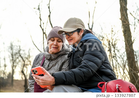 Two Russian women aged 50 and 60 sit together in a quiet park at dusk, calmly sharing a moment while looking at a phone and enjoying a sense of connection and peace. 133927807