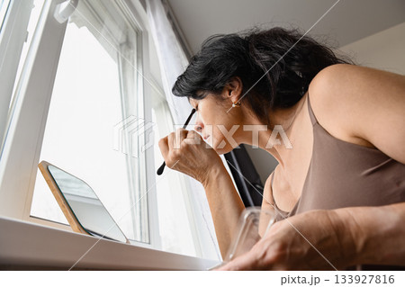 Russian mature woman doing eye makeup near the window showing mindful confidence soft emotion and a peaceful start to her day in natural light 133927816