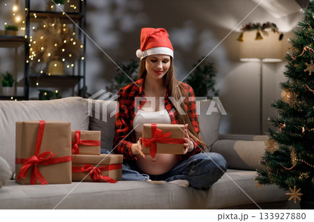 A pregnant woman wearing a Santa hat opens a Christmas gift with joy and anticipation A pregnant woman wearing a Santa hat opens a Christmas gift with joy and anticipation 133927880