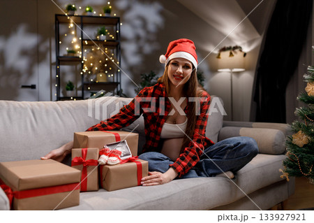 A pregnant woman wearing a Santa hat sits on a couch with wrapped gifts, celebrating the holiday season 133927921