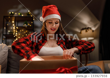 A woman wearing a Santa hat opens a gift box, anticipating the contents with excitement A woman wearing a Santa hat opens a gift box, anticipating the contents with excitement 133927938
