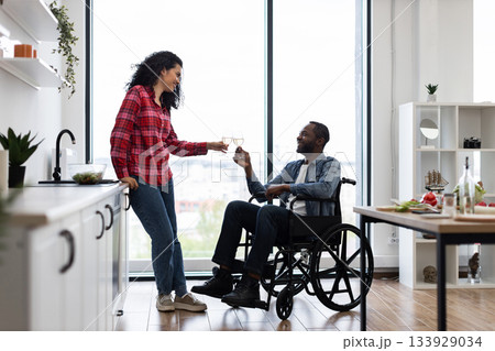 A diverse couple celebrates with wine in a bright, modern kitchen. One partner is in a wheelchair. A diverse couple celebrates with wine in a bright, modern kitchen. One partner is in a wheelchair. 133929034