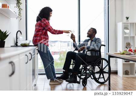 A woman offers a glass of wine to a man in a wheelchair, celebrating in a bright, modern kitchen setting. 133929047