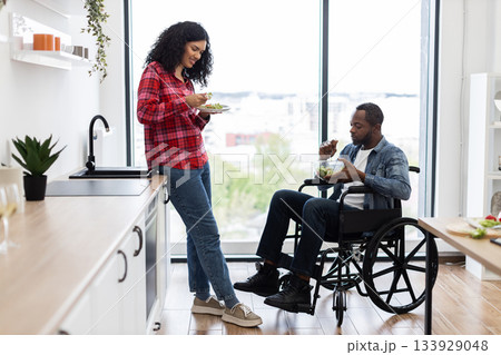 A diverse couple enjoys a healthy salad in a bright, modern kitchen. One person is in a wheelchair. 133929048