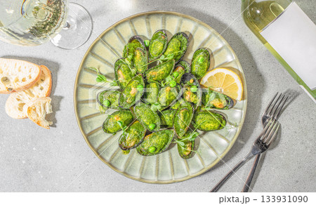 Gourmet baked mussels in green herb sauce served with lemon slices, white wine and bread on a light stone background, top view 133931090