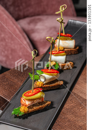Gourmet appetizer canapes with smoked meat, pickles, mustard seeds and herbs on dark rye bread served on a black platter in a fine dining restaurant Gourmet appetizer canapes with smoked meat, pickles, mustard seeds and herbs on dark rye bread served on a black platter in a fine dining restaurant 133931101