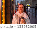 middle aged Caucasian woman drinks coffee in rain under neon lights. woman is walking alone in the city. 133932005