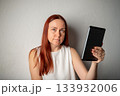 A Caucasian woman with natural beauty, no makeup, and long red hair holds a tablet device and looks intently at the camera. 133932006
