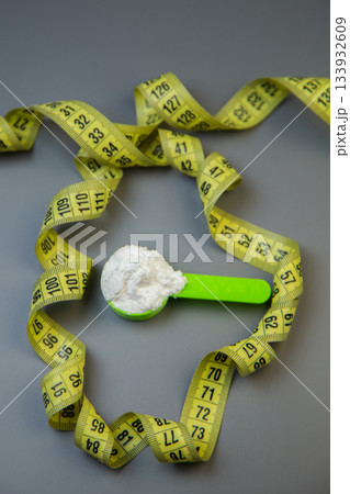 Green measuring spoon with white powder is surrounded by yellow measuring tape, representing health and fitness concepts, set against a gray background with copy space Green measuring spoon with white powder is surrounded by yellow measuring tape, representing health and fitness concepts, set against a gray background with copy space 133932609