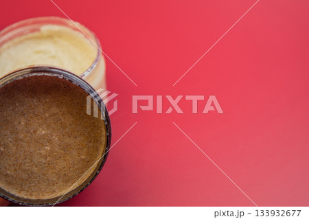 Natural body scrub jar reveals creamy brown texture, set against a vivid red backdrop, highlighting beauty, skincare, and self-care themes in a visually appealing manner 133932677