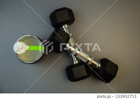 Black weights and a protein shake with a green spoon are arranged on a gray background, symbolizing fitness, strength training, and healthy nutrition choices 133932713