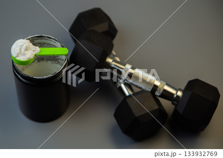 Set of black dumbbells alongside a protein powder container with a green scoop, placed on a gray surface, emphasizing fitness and nutrition essentials for workouts 133932769