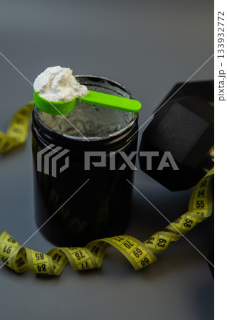 Black protein powder container features green scoop on top, surrounded by yellow measuring tape and dumbbells, representing fitness and health goals 133932772