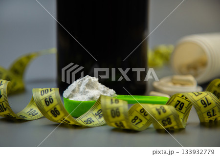 Measuring spoon with white powder on gray surface, encircled by yellow measuring tape and dark bottle, illustrating health, nutrition, and fitness themes Measuring spoon with white powder on gray surface, encircled by yellow measuring tape and dark bottle, illustrating health, nutrition, and fitness themes 133932779